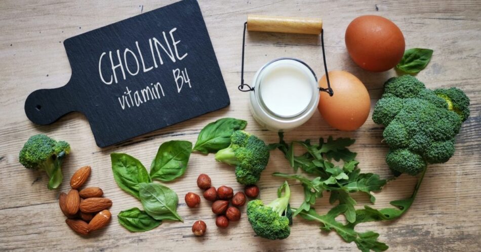 Na drewnianym stole leżą różne produkty spożywcze bogate w cholinę. Po lewej stronie znajduje się tabliczka z napisem "CHOLINE vitamin B4". Wokół niej rozmieszczone są brokuły, świeże liście szpinaku i rukoli, migdały, orzechy laskowe, dwa jajka oraz szklanka mleka z drewnianym uchwytem. Produkty te symbolizują naturalne źródła choliny w diecie.