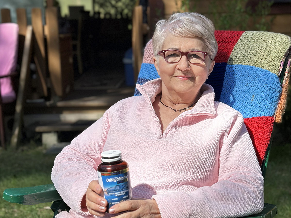 Małe kapsułki bez posmaku tranu Pani Barbara siedzi na kolorowym leżaku na działce i trzma opakowanie Omega Marine.