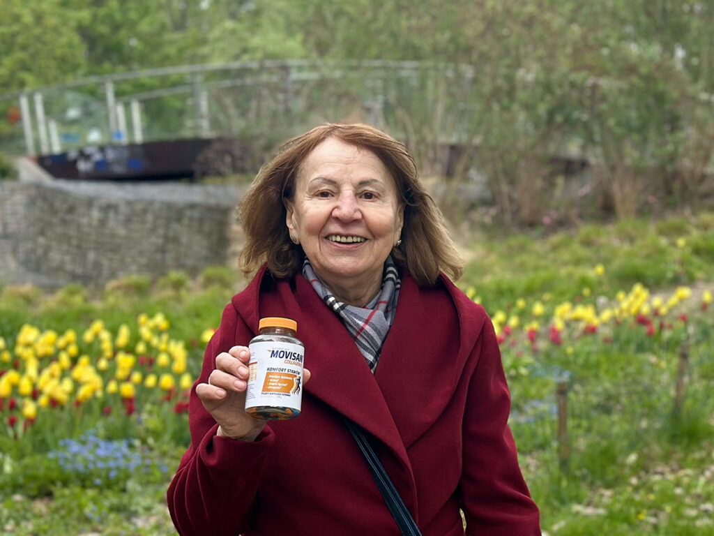 Pani Lucyna stoi na tle żółtych tulipanów w czerwonym płaszczu, trzymając opakownie Movisanu.